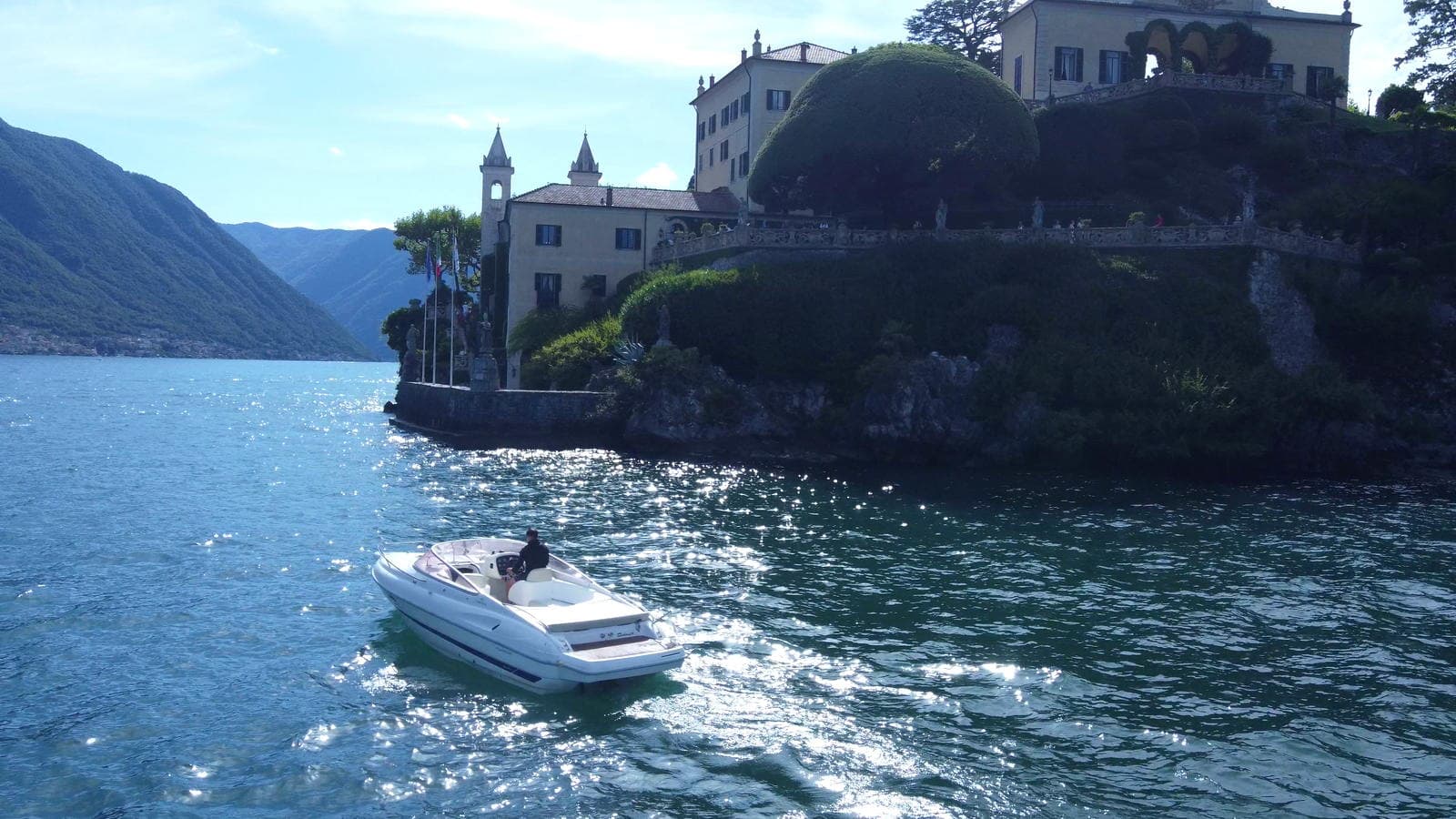 Villa del Balbianello seen close from the water, afternoon light scattering across the lake as the boat passes below