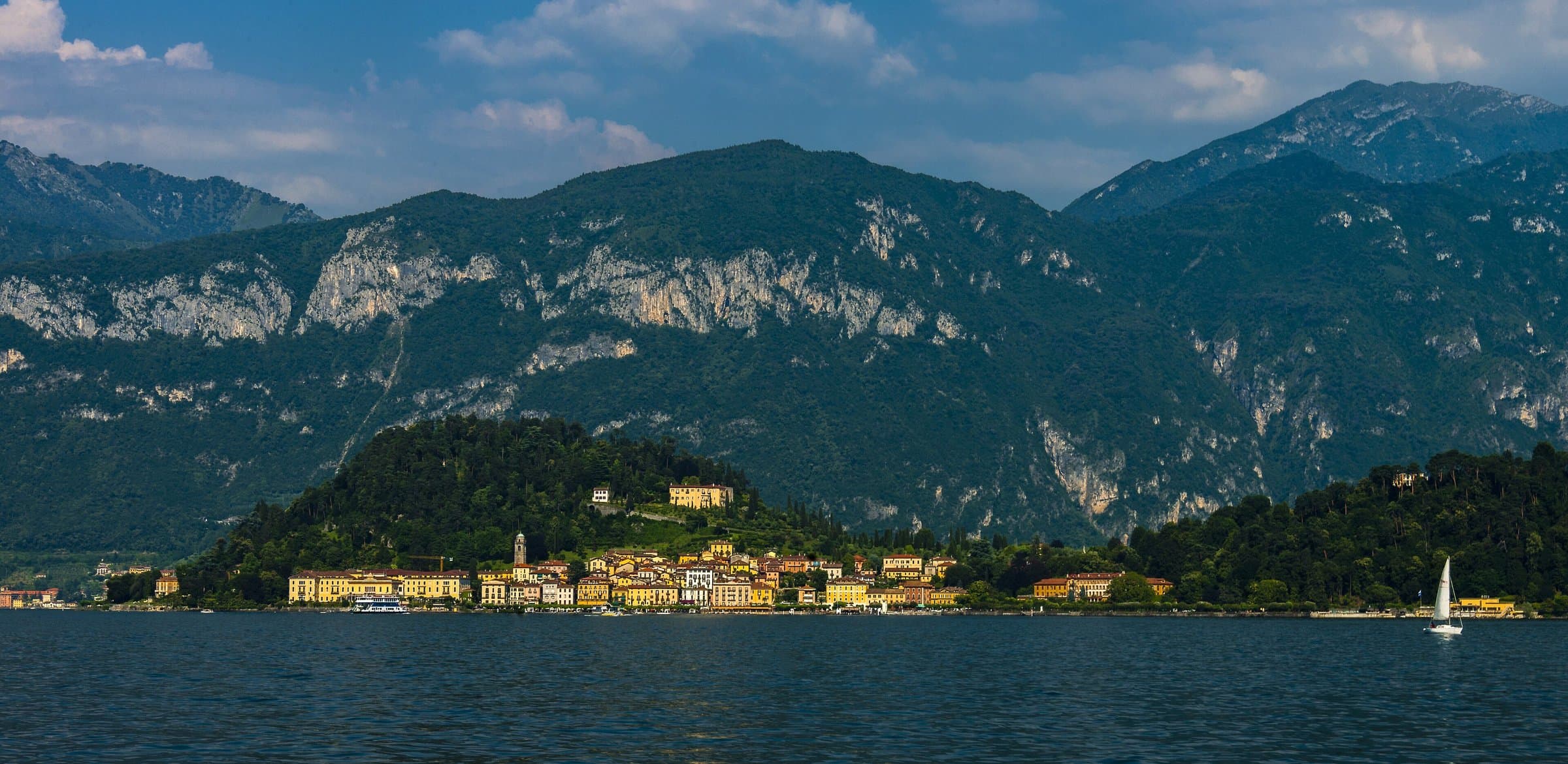Bellagio glowing in the sun at the foot of the Grigna mountains, seen from the water of Lake Como with a small sailboat passing
