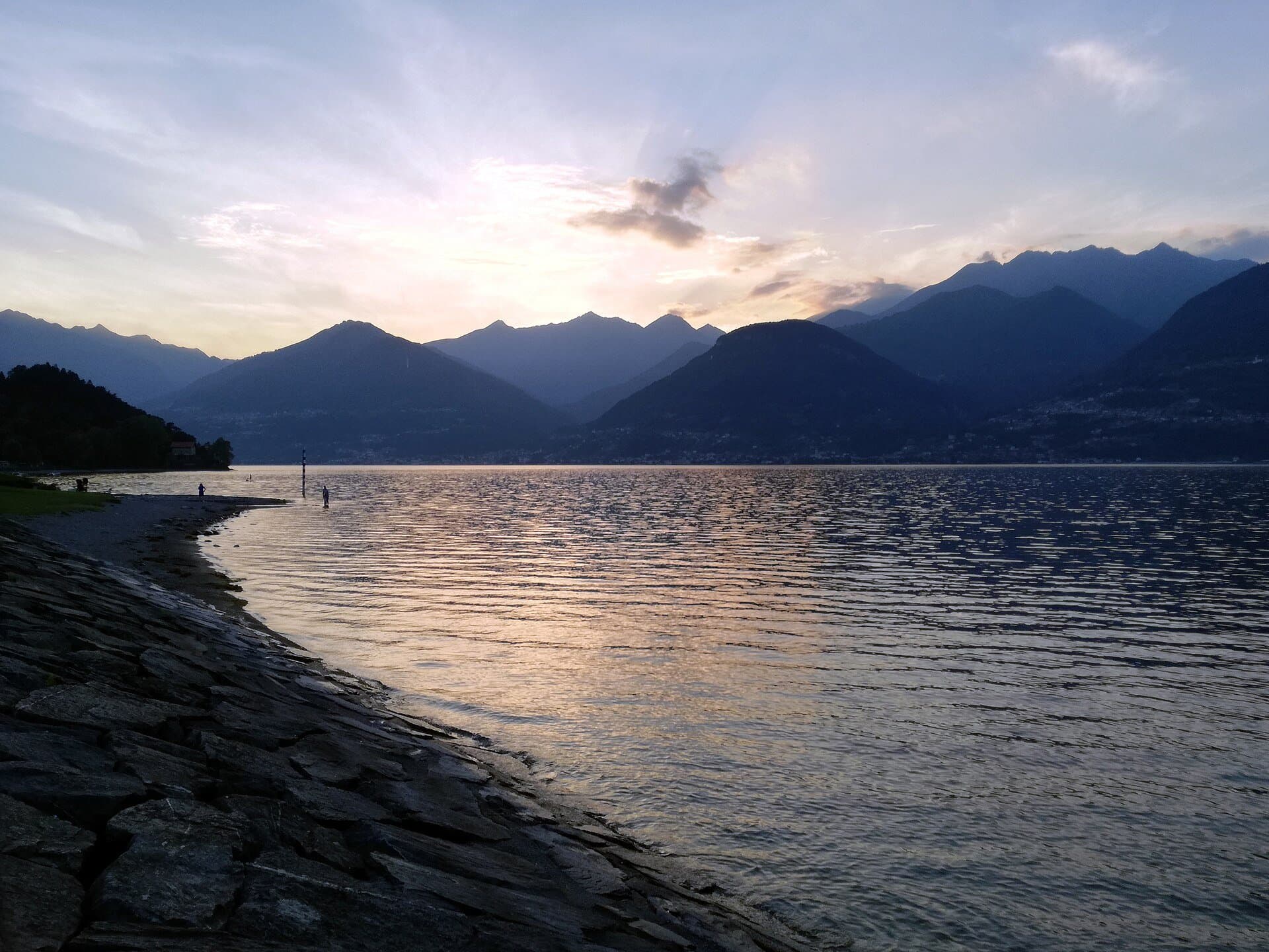 Sunset over Lake Como from a stone shore, with the mountains of the western branch fading into golden light