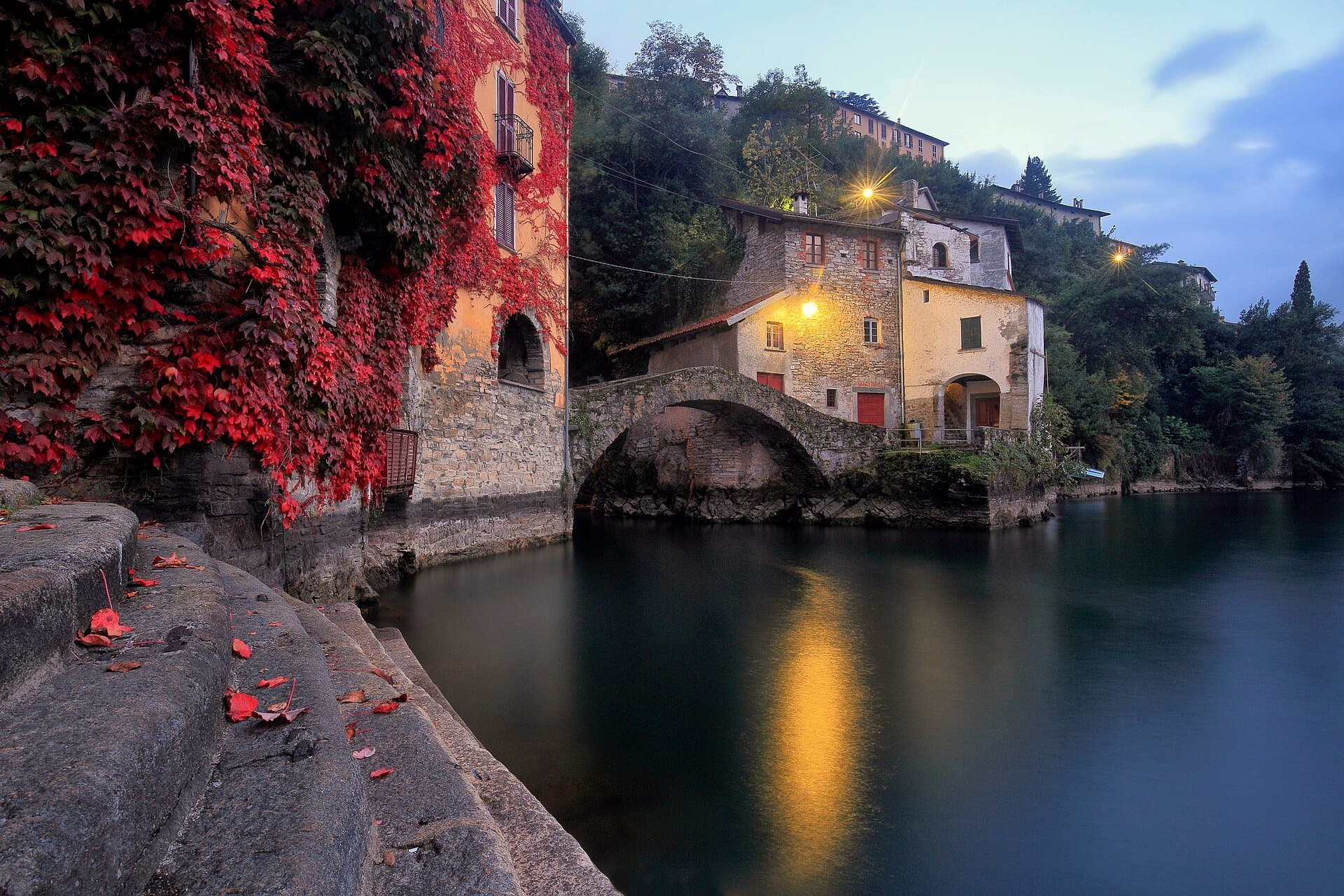 The medieval Ponte della Civera stone bridge in Nesso at dusk, framed by red autumn ivy and reflected in the green water of Lake Como