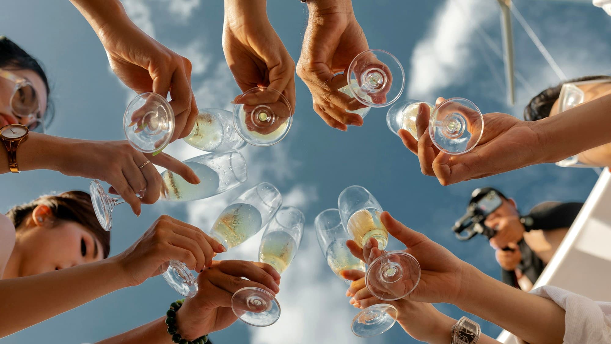Group of friends raising champagne flutes together against a blue summer sky, toasting on a boat
